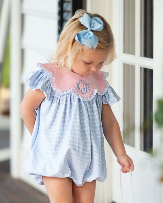 Child wearing a light blue dress with a pink collar and monogram, standing indoors.