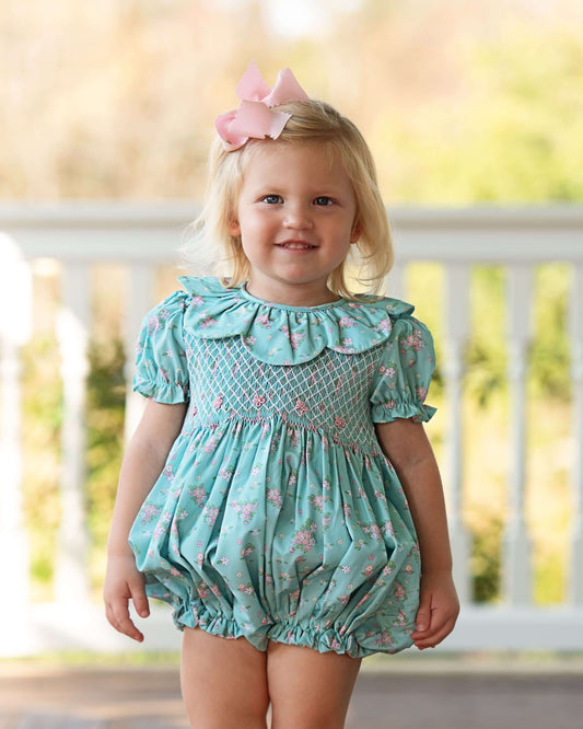 Child wearing a light blue floral romper with a pink bow in hair, standing on a wooden deck.