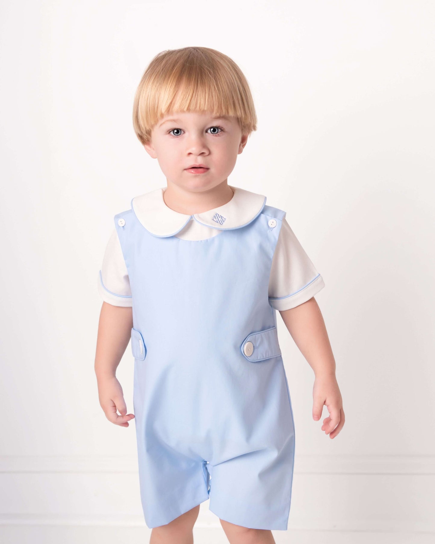 Child wearing a light blue romper with a white shirt underneath on a plain background