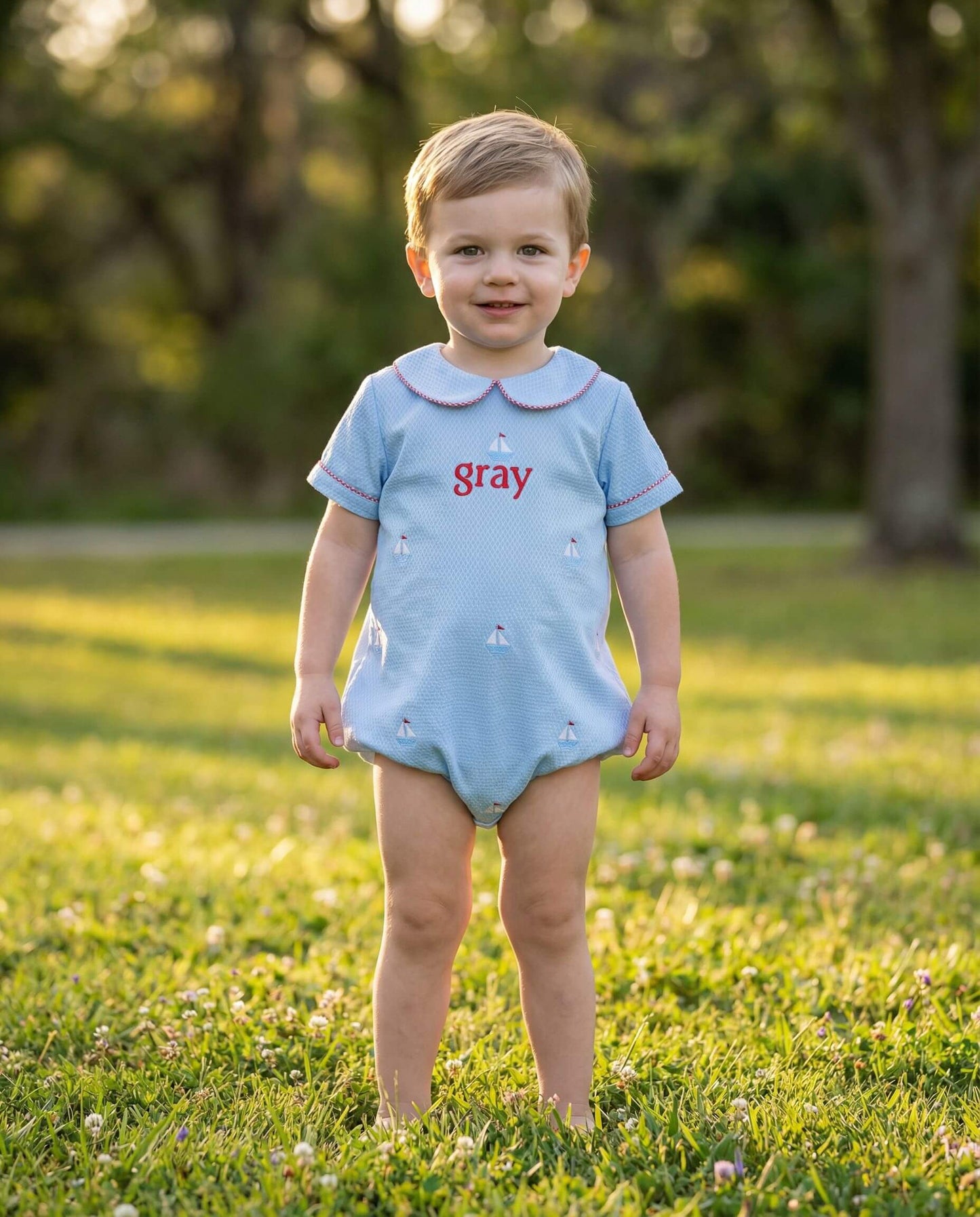 Child wearing a light blue romper with 'gray' printed on it, standing in a grassy outdoor setting.