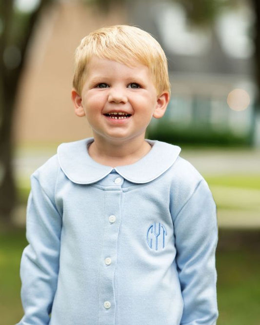 Child wearing a light blue shirt with a collar and monogram, standing outdoors.