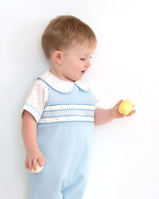 Child wearing a light blue smocked dress over a white shirt, holding a yellow toy against a white background