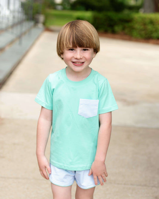 Child wearing a light blue t-shirt with a pocket and striped shorts, standing on a pathway.