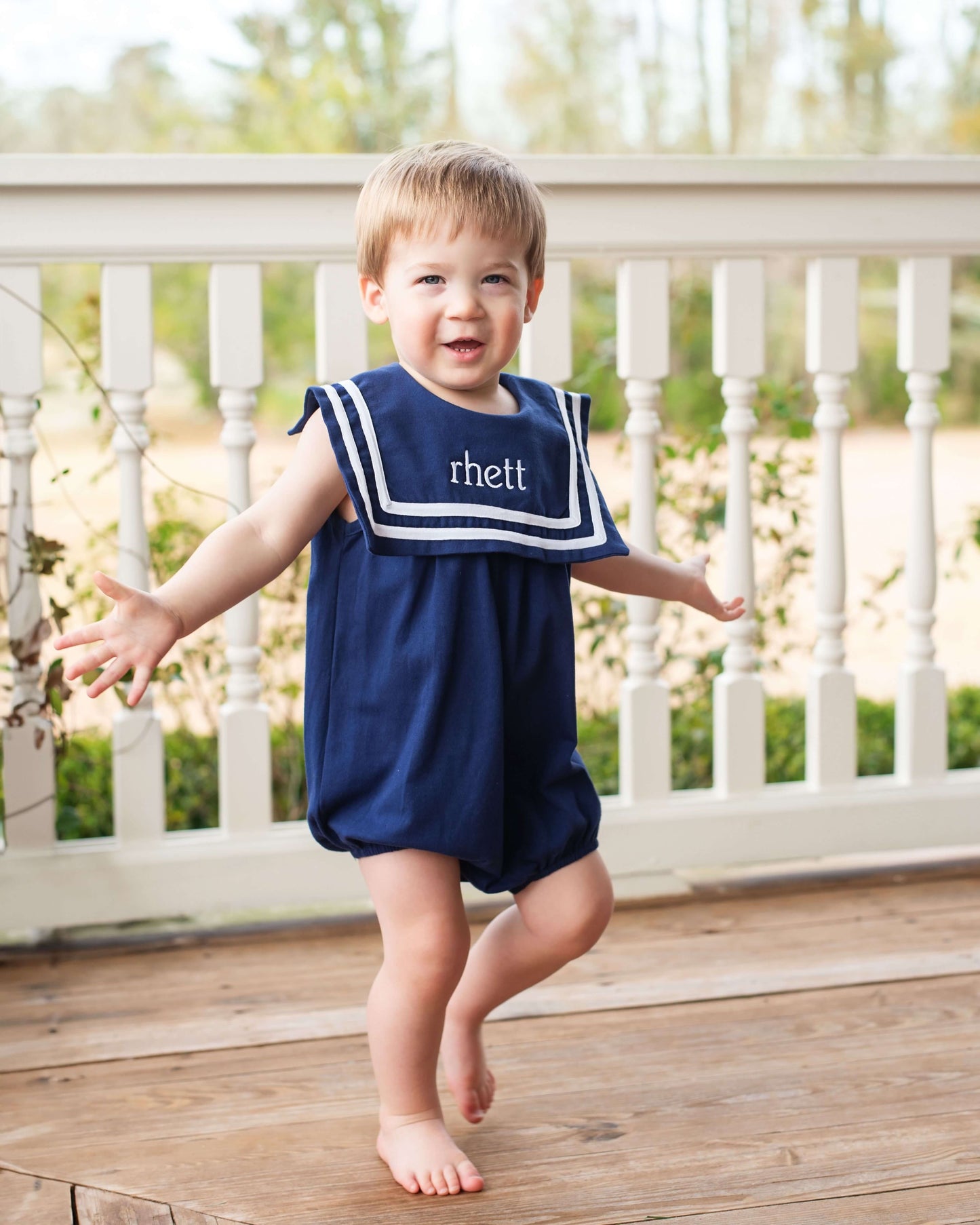 Child wearing a navy blue romper with 'rhett' on a wooden deck