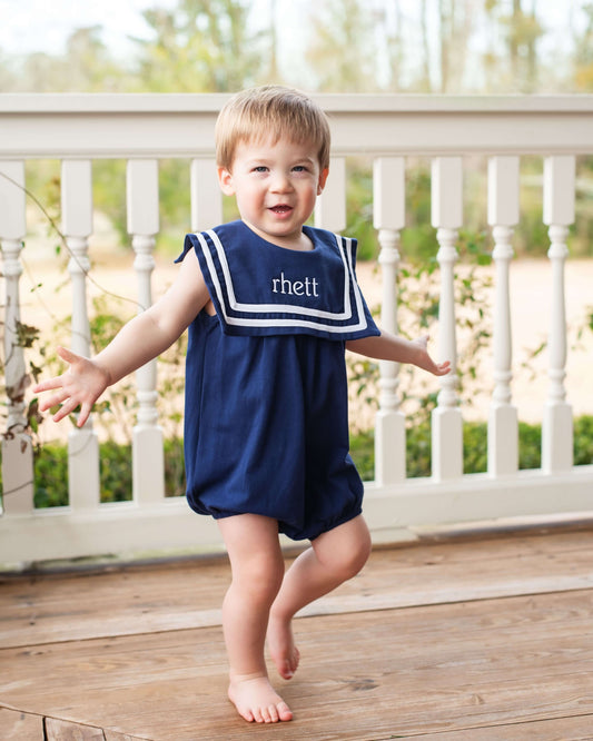 Child wearing a navy blue romper with 'rhett' on a wooden deck