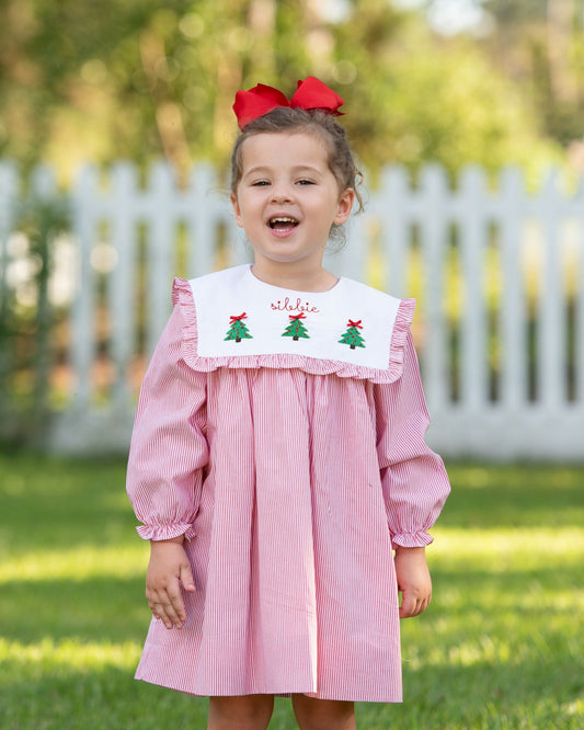 Child wearing a pink dress with green tree designs and red bows outdoors.