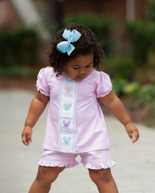 Child wearing a pink outfit with decorative buttons and a blue bow in hair, standing outdoors.