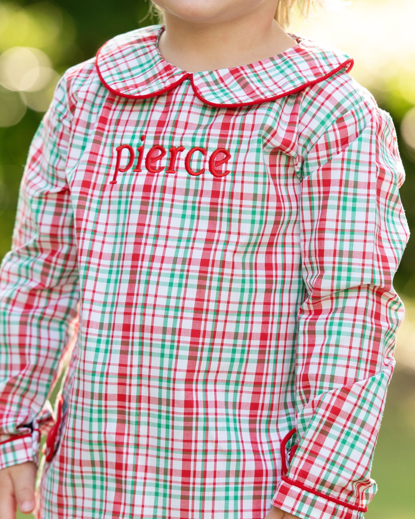 Child wearing a red and green checkered dress with 'pierce' embroidered on it, outdoors.