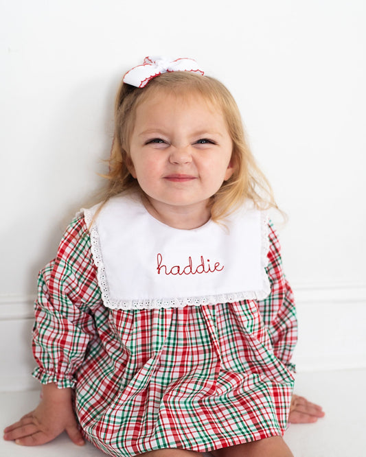 Child wearing a red and green checkered outfit with a white bib and embroidered name on a white background