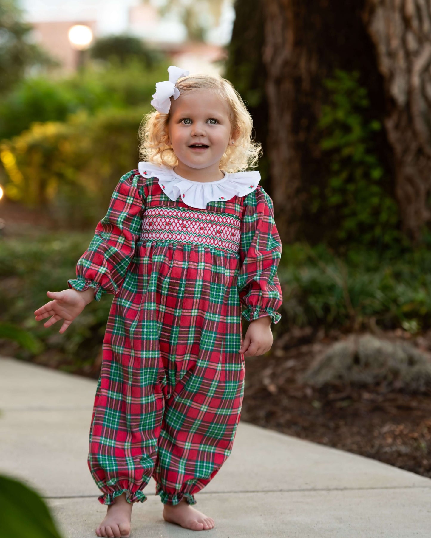 Child wearing a red and green plaid outfit with a white bow outdoors.