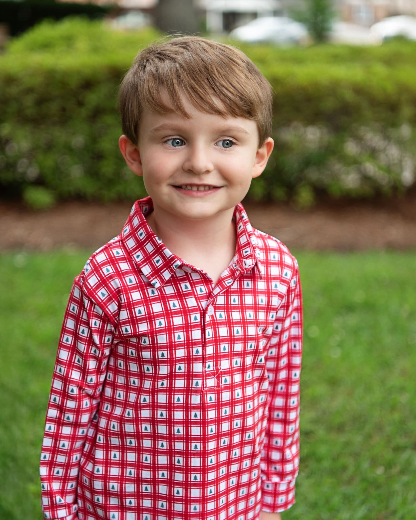 Child wearing a red checkered shirt standing outdoors with greenery in the background