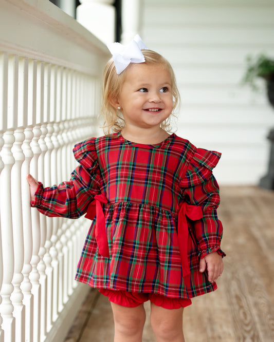 Child wearing a red plaid dress with ruffles standing on a wooden floor.