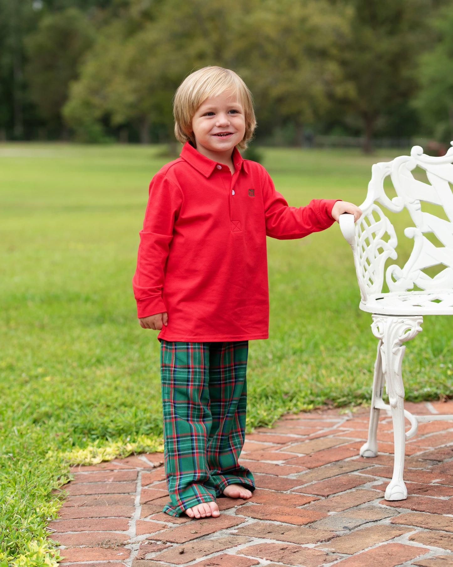 Child wearing a red shirt and plaid pants standing on a brick path with green grass and trees in the background.