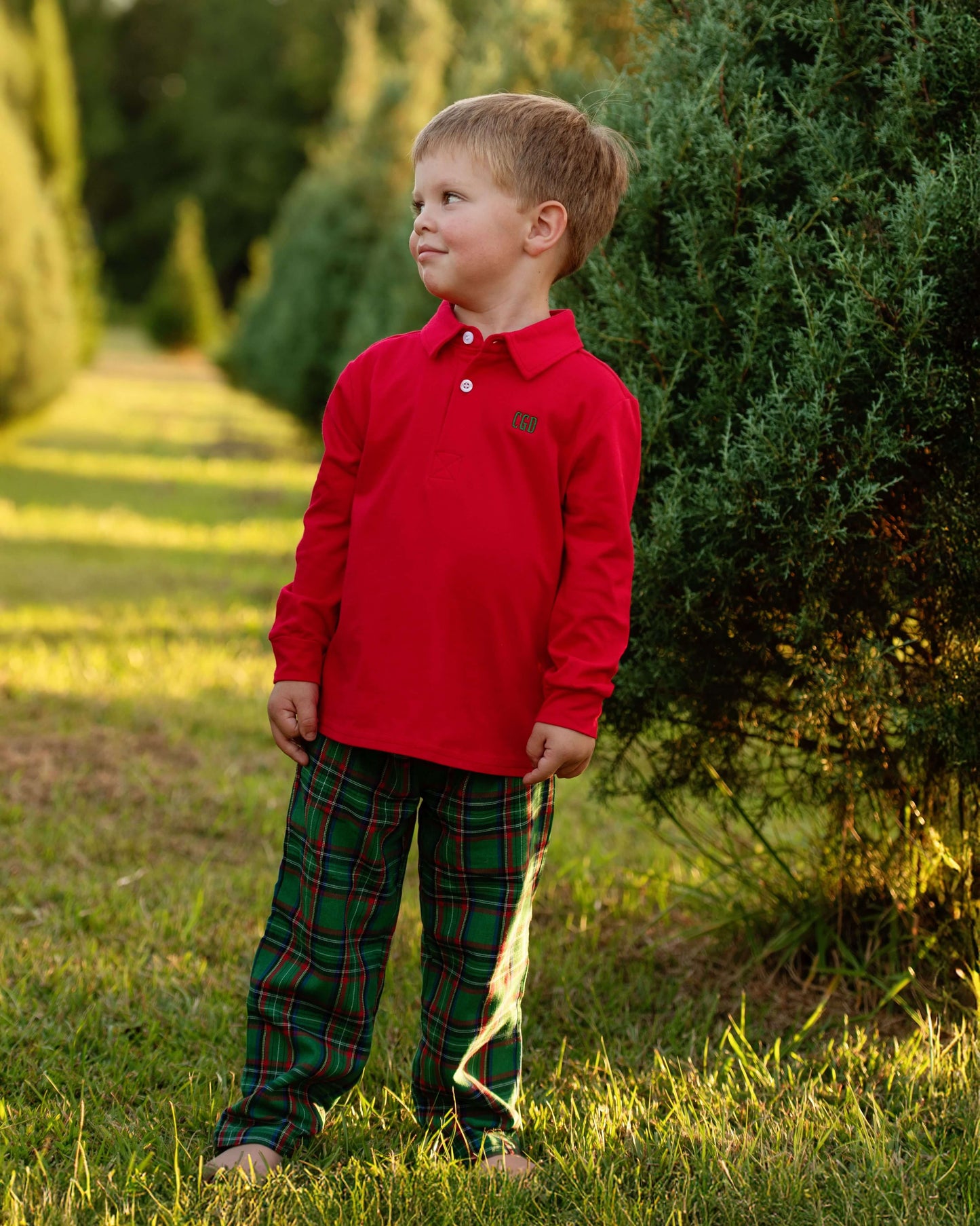 Child wearing a red shirt and plaid pants standing outdoors near greenery.
