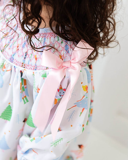 Child wearing a smocked long bubble with a Christmas print and a large pink bow on a plain background