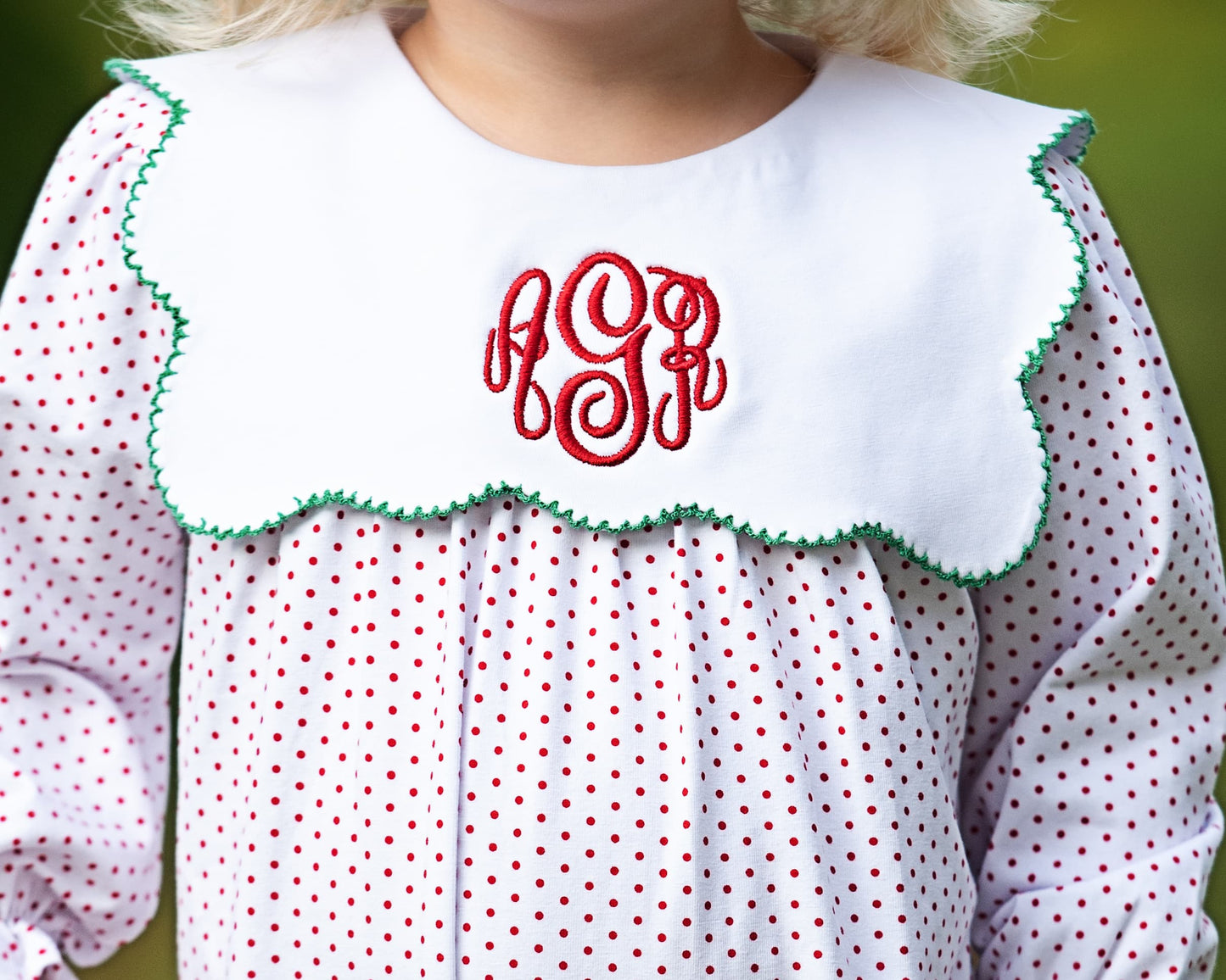 Child wearing a white blouse with red polka dots and green trim, featuring a red monogram.