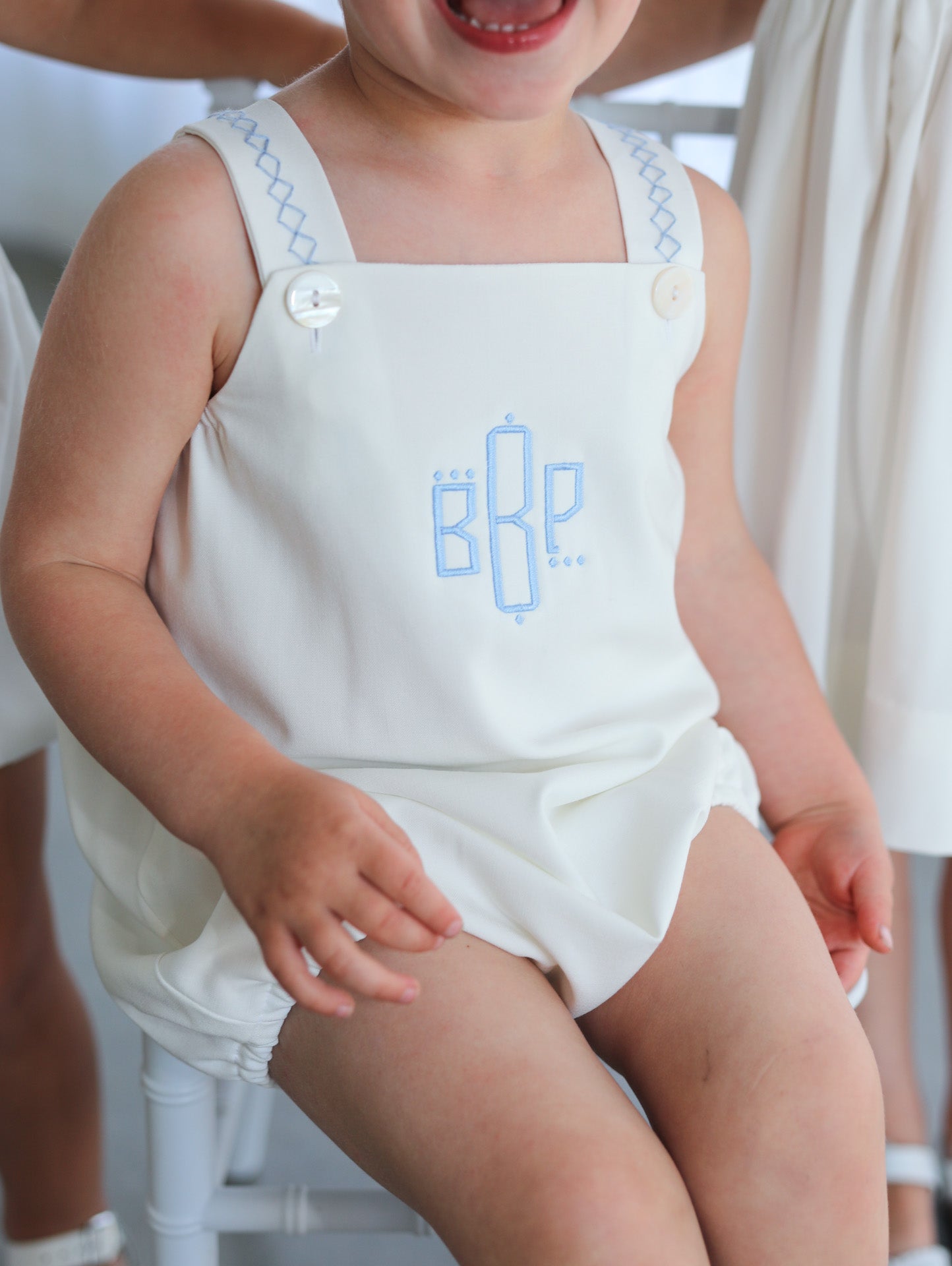 Child wearing a white bubble with blue embroidery, sitting on a white chair.