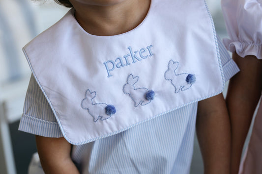 Child wearing a white collar with embroidered name and bunnies, standing against a neutral background.