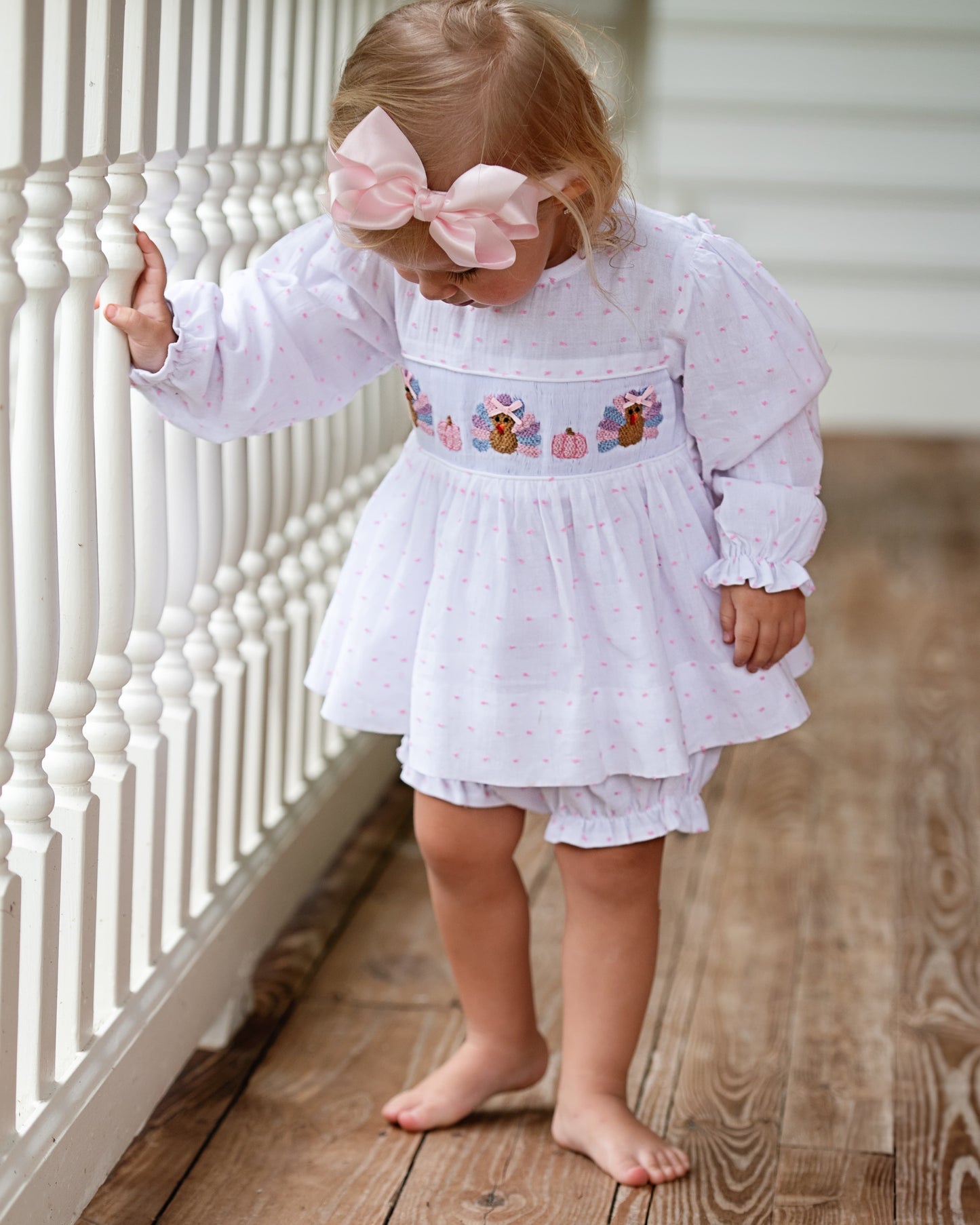 Young child wearing a white dress with embroidery, standing on a wooden floor.