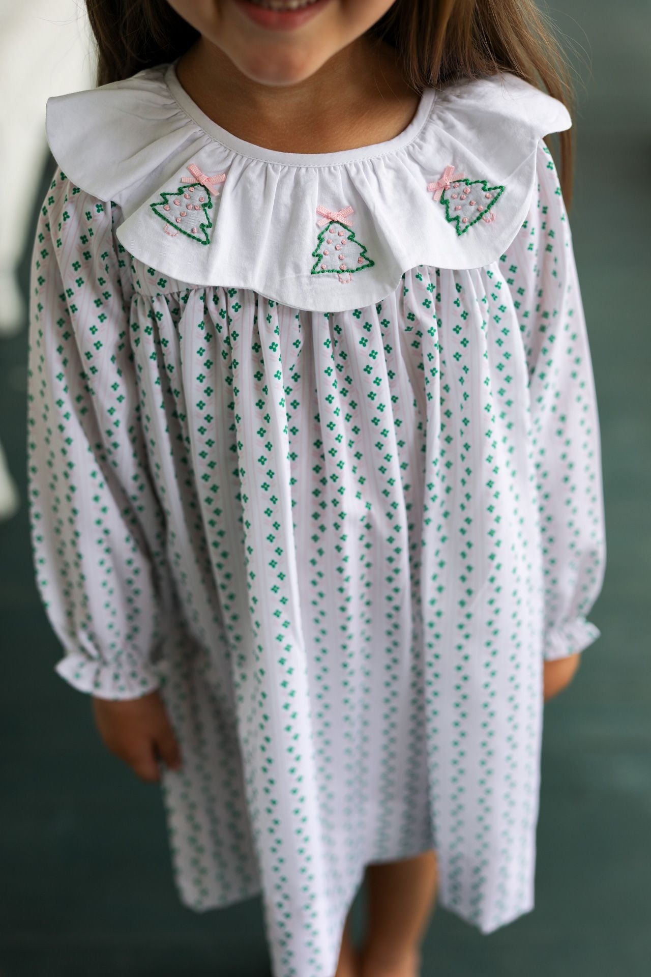 Child wearing a white dress with green patterns and a white collar against a neutral background