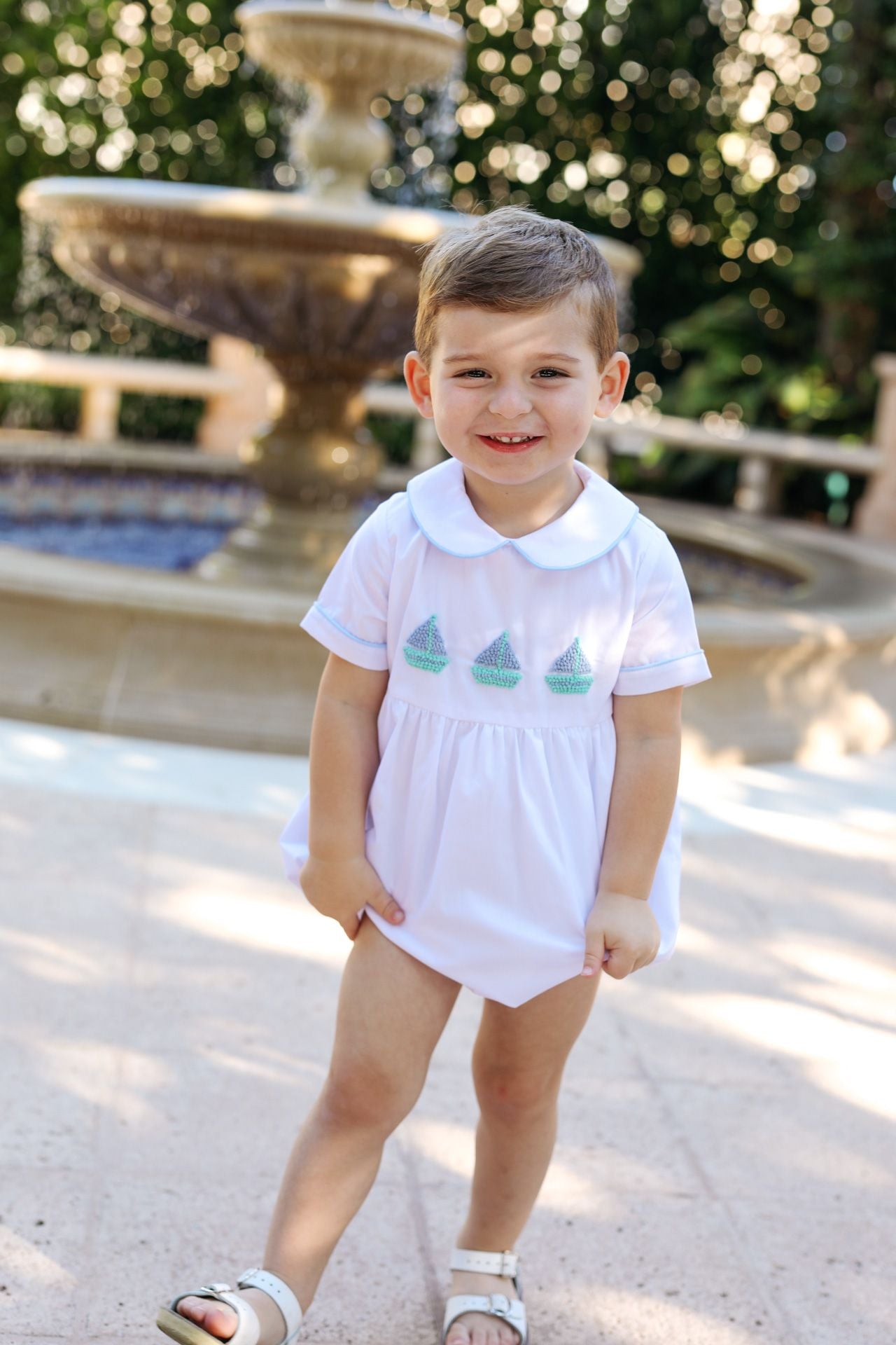 Child wearing a white outfit with green sailboat designs in front of a fountain.