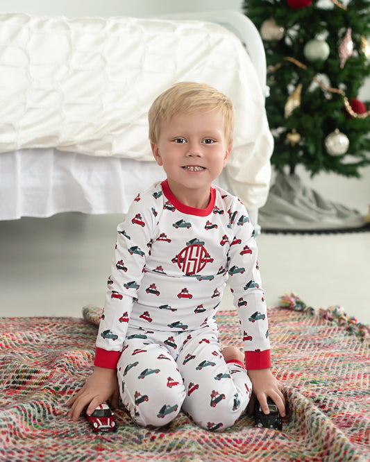 Child wearing pajamas with car pattern sitting on a rug in front of a Christmas tree.