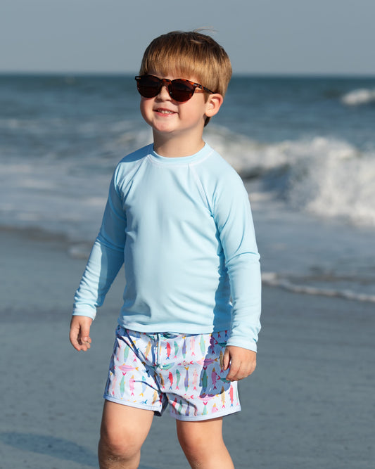 Child wearing sunglasses and a light blue long-sleeve shirt on a beach.