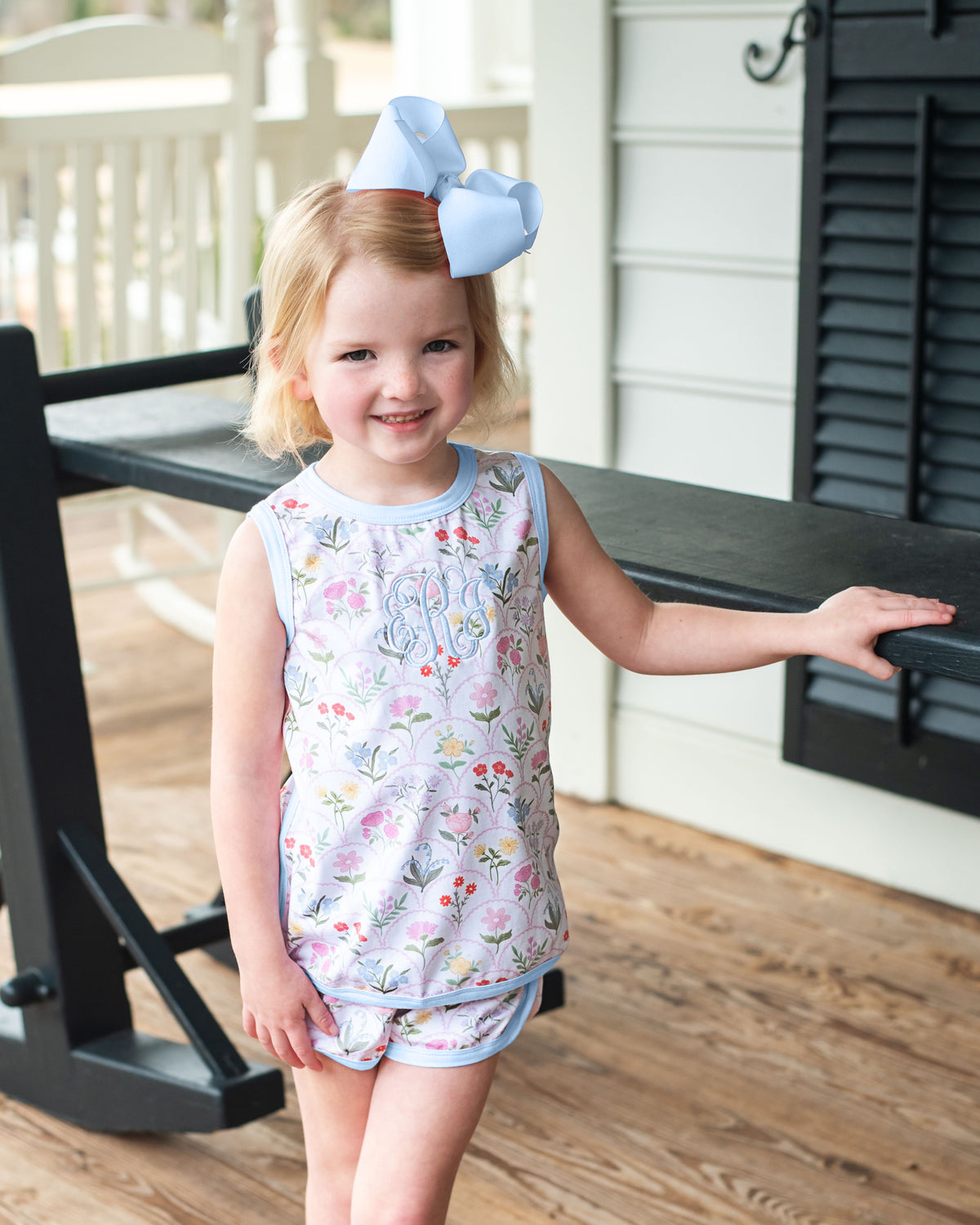 Young girl in a floral outfit with a large blue bow standing on a wooden porch.