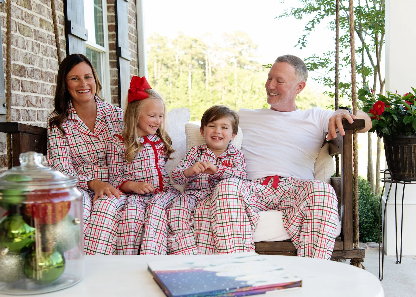 Family of four wearing matching red and white plaid pajamas sitting on a porch.