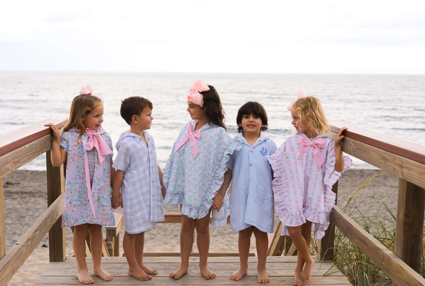 Five children in swimwear and coverups standing on a wooden deck by the beach.