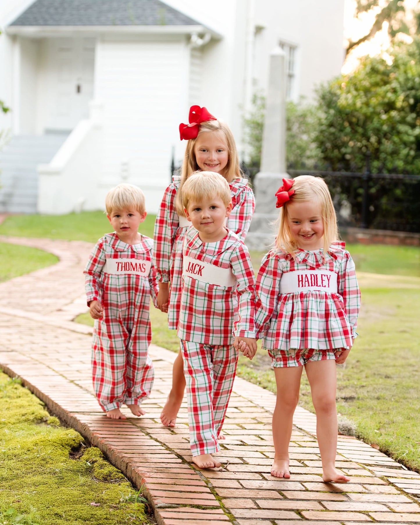 Four children in matching red and white checkered outfits standing on a wooden path outdoors.