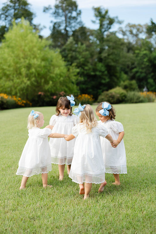 Four young girls in white dresses with blue bows standing in a circle on a grassy field.