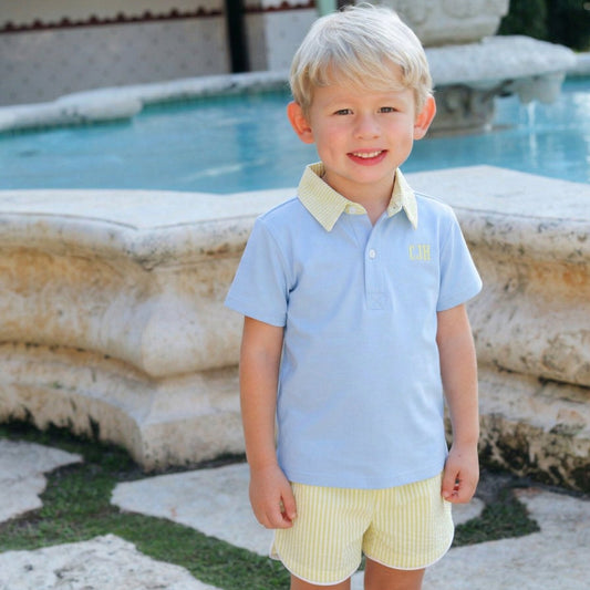 Young boy in a light blue shirt and yellow shorts standing in front of a fountain.