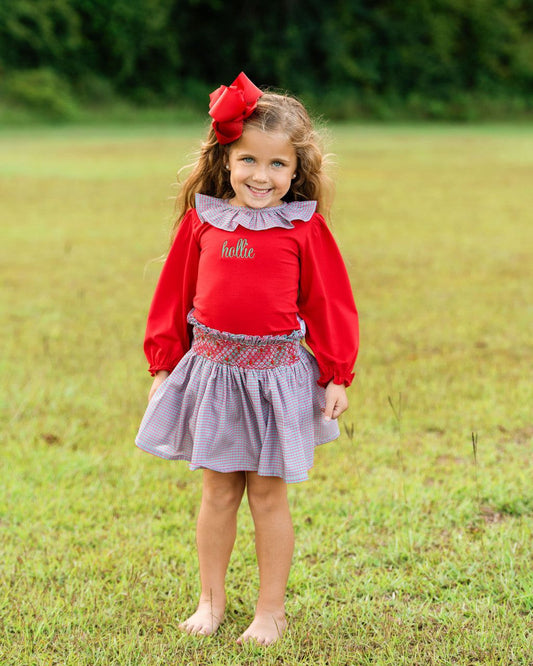 Girl standing on the grass barefoot wearing a red and plaid outfit with a red bow on her hair
