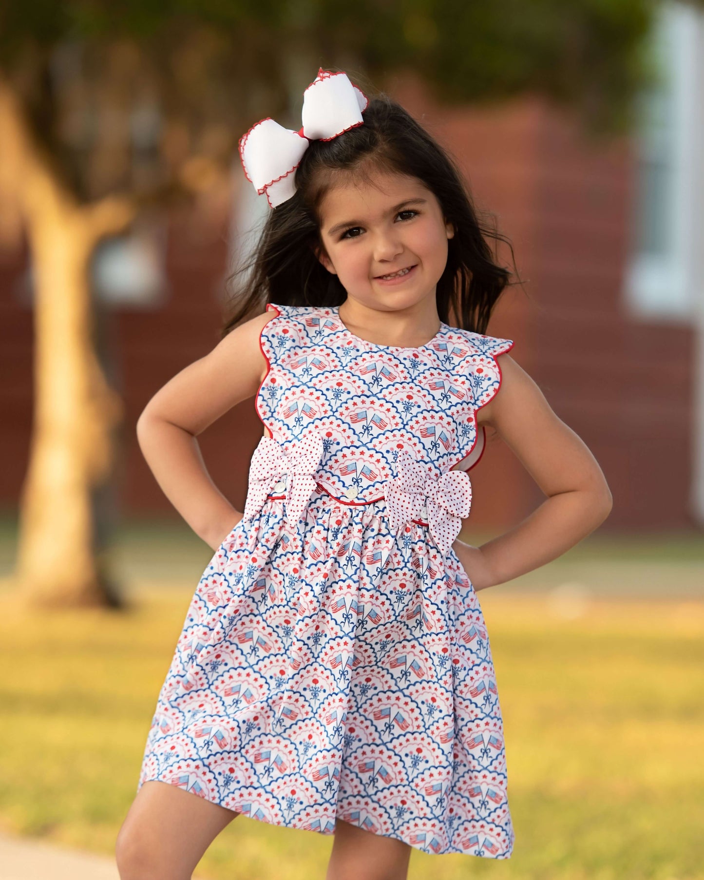 Girl wearing a flag print dress with a white bow on her hair and hands on her hips.