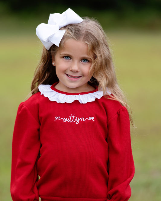 Girl with blonde hair wearing a red sweater with embroidery and a white bow on her hair