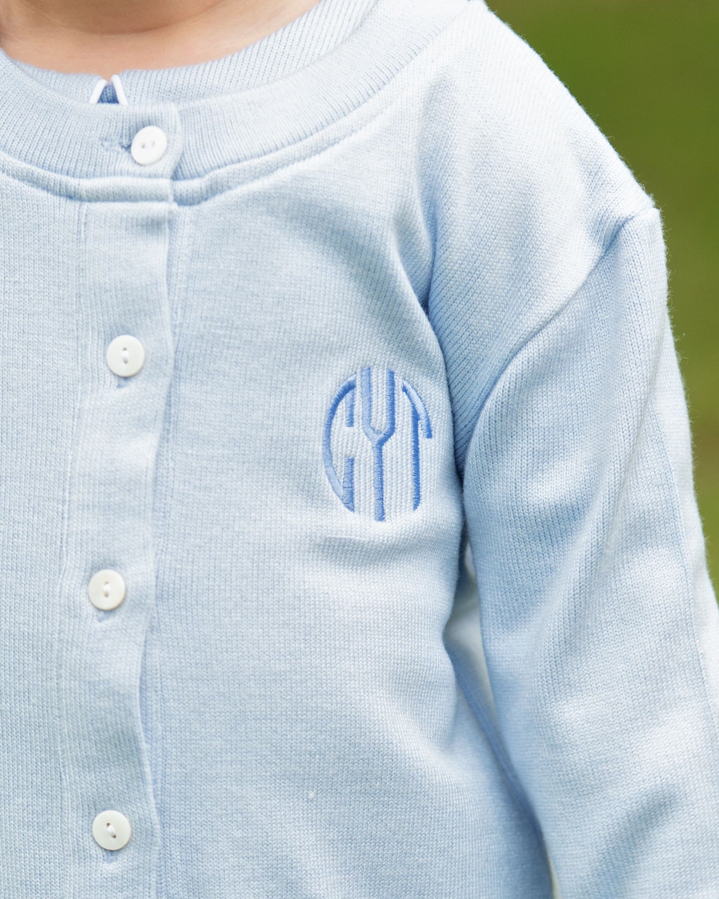 Light blue cardigan with monogram on a blurred green background