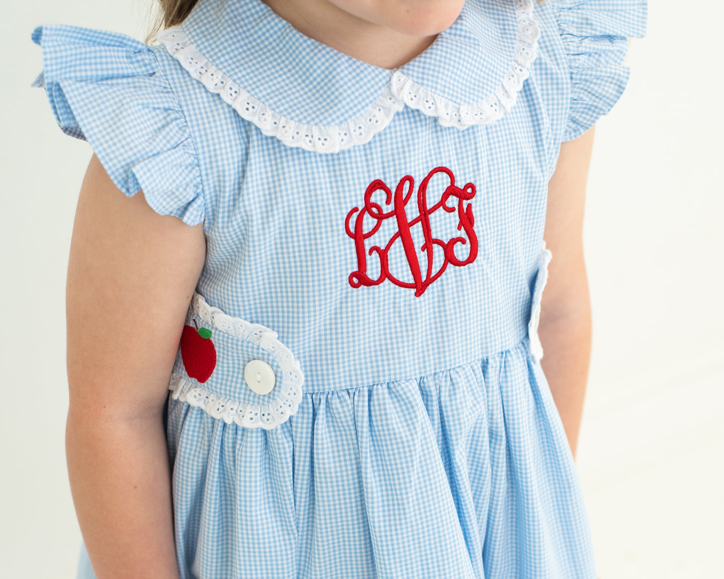 Light blue checkered dress with white lace collar and red monogram on a plain background