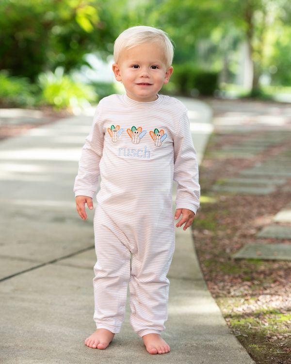 Little boy in a khaki long romper with turkey and monogram "rusch", standing on a path outdoors.