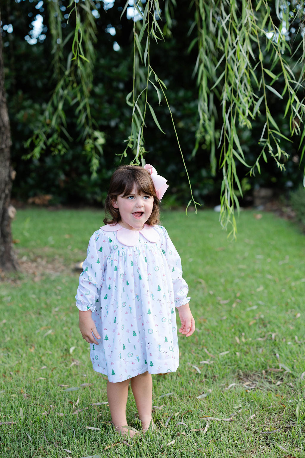 Little girl standing in the grass wearing a Christmas dress and a pink bow in her hair.
