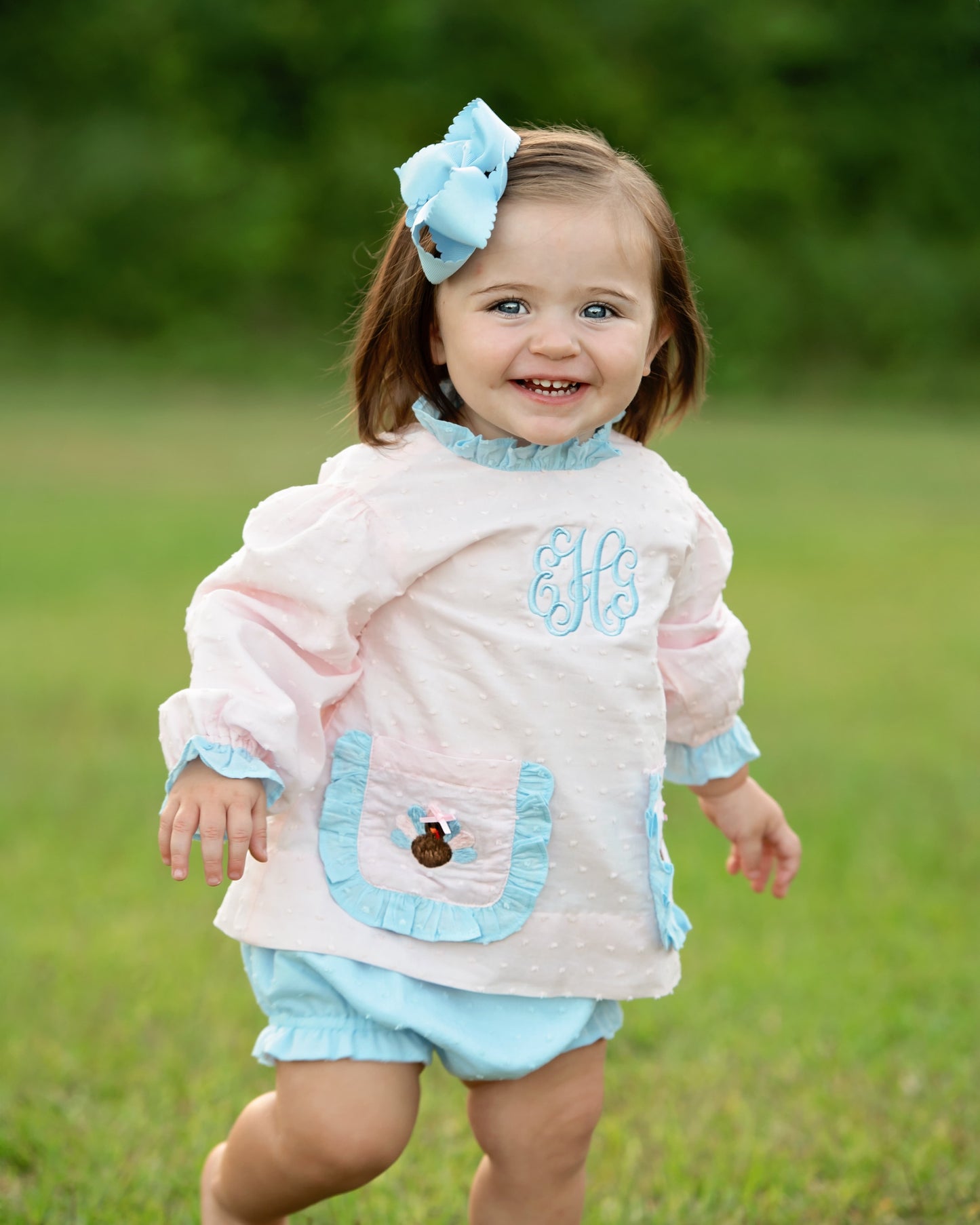 Little girl wearing a pink outfit with turkeys and a bow, standing in a grassy field.