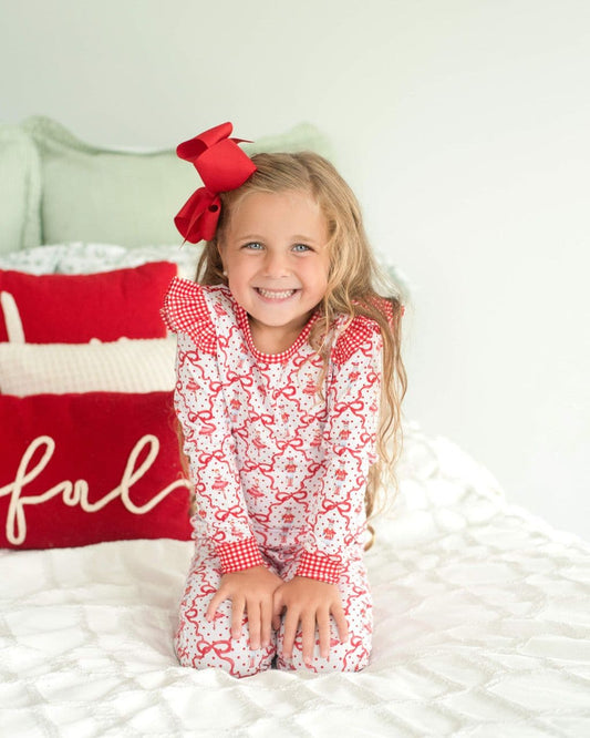 Little girl wearing holiday pajamas with ballerinas, sitting on a bed with a red bow in her hair