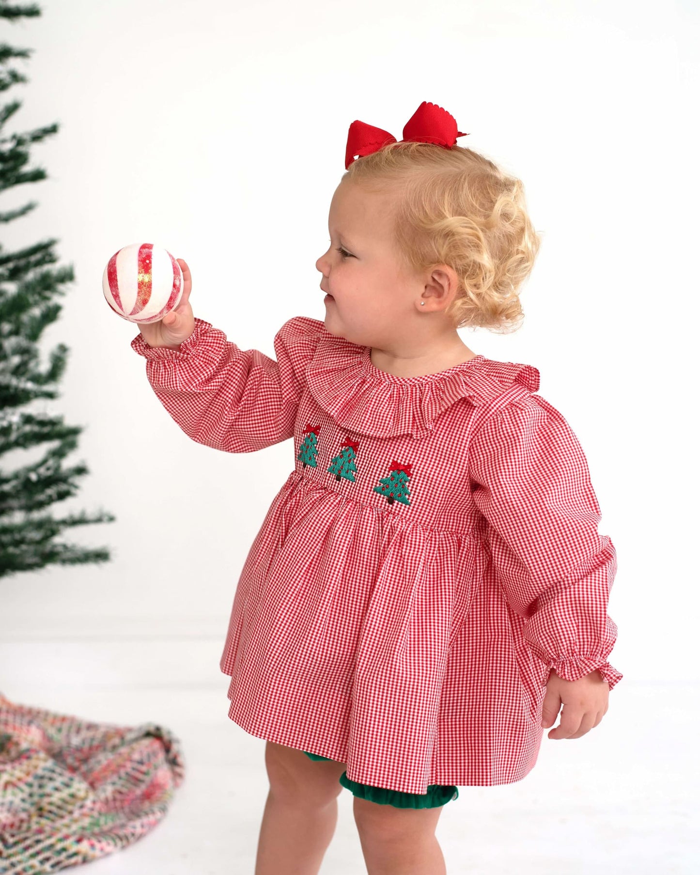 A toddler girl wearing a red and white gingham long-sleeve dress with embroidered Christmas trees on the chest and a red bow in her hair, holding a striped ornament against a white background with a Christmas tree on the left.