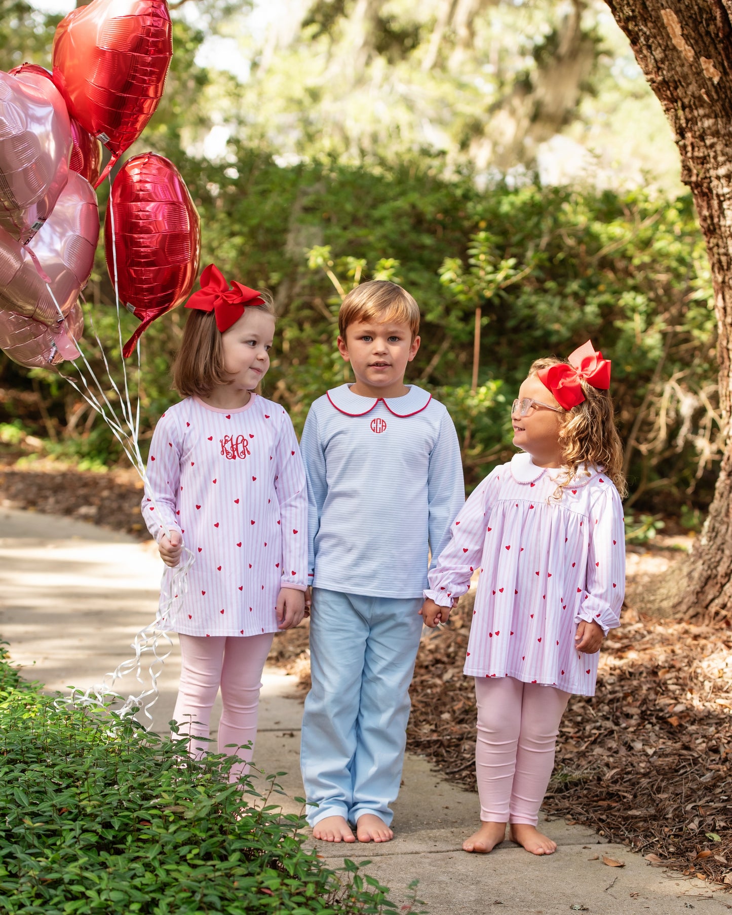 Three children in Valentine outfits with red bows outdoors, holding balloons.