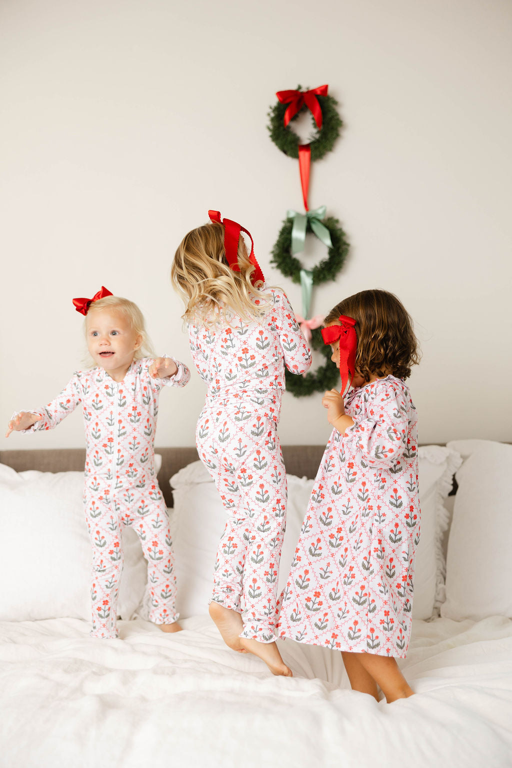 Three children in matching pajamas standing on a bed with Christmas wreaths in the background.
