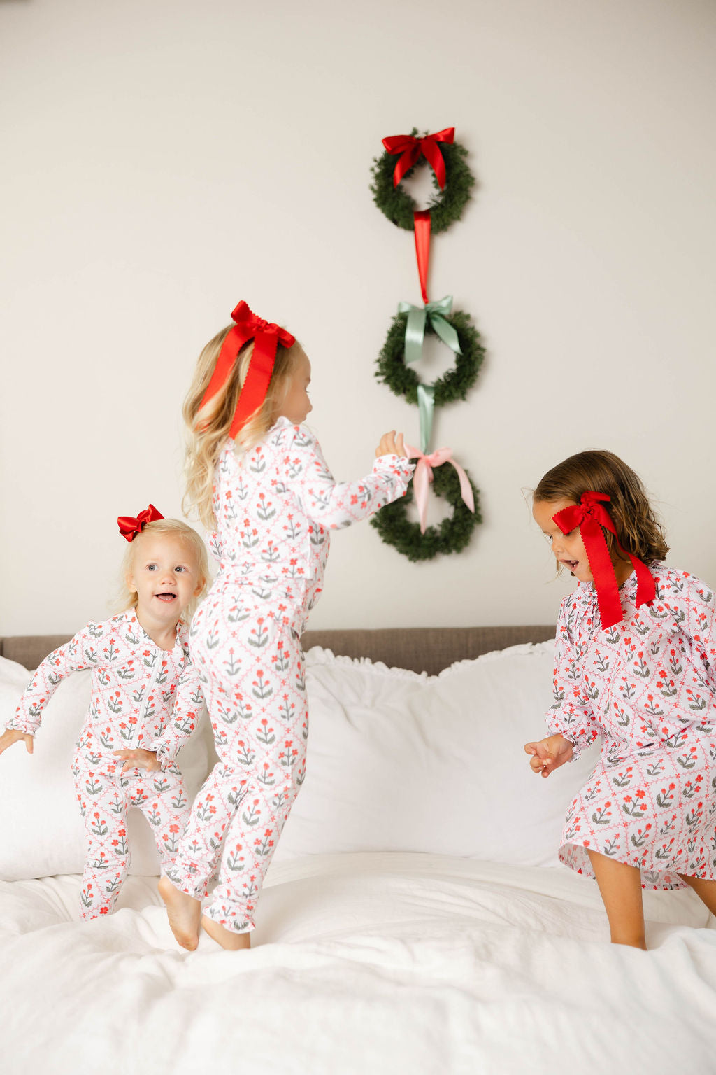 Three children in matching pajamas with red bows standing on a bed with Christmas wreaths hanging above.