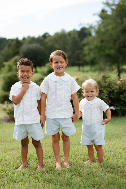 Three children in matching white outfits standing outdoors with greenery in the background