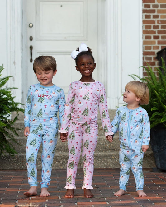 Three children standing barefoot on a brick porch wearing matching long-sleeve pajamas with a Christmas tree print. The child on the left wears a light blue pajama set with 'lil' boy' printed on the chest, the center child wears a pink pajama set with a red and green tree print, and the child on the right wears a matching light blue pajama set.