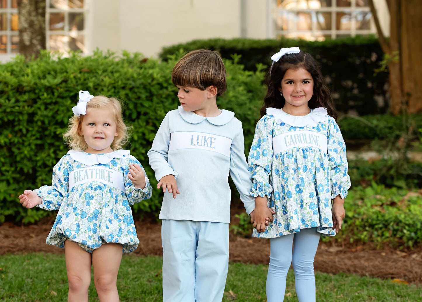 Three children wearing matching outfits with 'LUKE' and floral patterns outdoors.