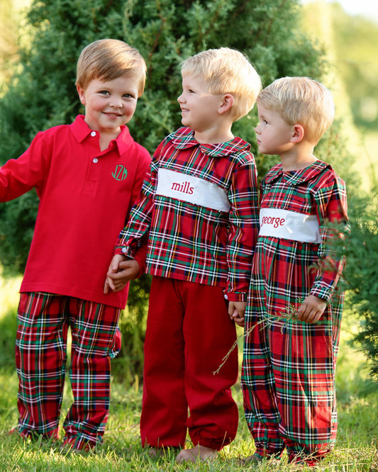 Three young boys standing outdoors in a grassy area wearing matching holiday-themed pajamas. Two boys wear red plaid long-sleeve pajamas with white name patches, and one boy wears a solid red long-sleeve pajama shirt with matching red plaid pants.